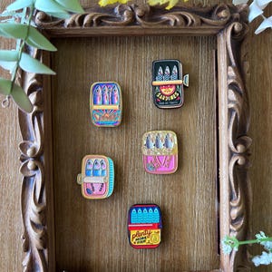 May include: Five rectangular enamel pins shaped like sardine tins, each featuring three fish illustrations. The pins display colorful designs with text including SARDINES, POUR LES TOASTS, IT'S FINE, and PLENTY MORE FISH, arranged inside a decorative wooden frame.