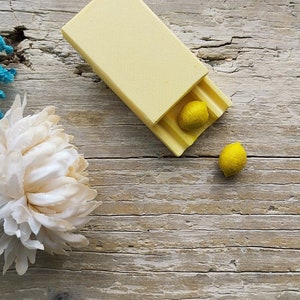 Può includere: Una scatola di legno gialla con un coperchio incernierato, aperta per rivelare due piccole sfere gialle all'interno. La scatola è appoggiata su una superficie di legno marrone chiaro.