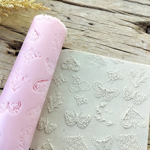 Könnte beinhalten: Eine rosa Teigrolle und eine grau strukturierte Oberfläche mit geprägten Schmetterlings- und Blumendesigns. Die Teigrolle zeigt ein sich wiederholendes Muster aus Motten, Blumen und Monden. Die Oberfläche zeigt eine Vielzahl detaillierter Motten- und Blumendesigns.