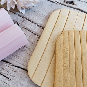 May include: Two silicone baking mats with a wood grain texture. The larger mat is light brown and the smaller mat is a darker brown.
