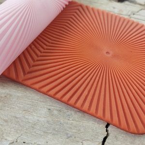 Puede incluir: Primer plano de una estera de hornear de silicona roja con un diseño de rayos de sol. La estera se está enrollando con un rodillo de silicona rosa.