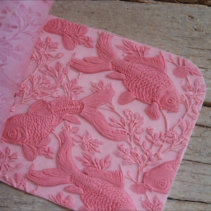 Puede incluir: Molde de silicona rosa rectangular con un diseño en relieve detallado de peces koi nadando entre plantas acuáticas. Los peces tienen escamas texturizadas y aletas fluidas, creando un efecto tridimensional. Para manualidades.