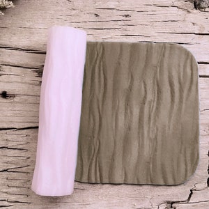 May include: A light pink silicone rolling pin with a wood grain texture. The rolling pin is rolled out on a brown textured surface.