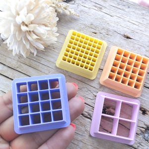 May include: Four plastic square-shaped food cutters with different grid patterns. The cutters are purple, yellow, orange, and pink.