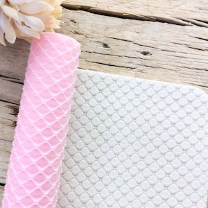 May include: A rolled pink silicone mat with a diamond pattern sits next to a white silicone mat with the same pattern. The mats are on a weathered wooden surface. A light pink flower is in the upper left corner.