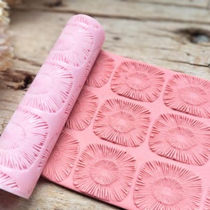 Könnte beinhalten: Ein rosa Silikon-Nudelholz- und Matten-Set. Das Nudelholz ist aufgerollt und zeigt ein sich wiederholendes Muster aus quadratischen Formen mit einem Sonnenstrahl-Design. Die Matte ist flach und zeigt das gleiche Muster. Für Backen und Basteln.
