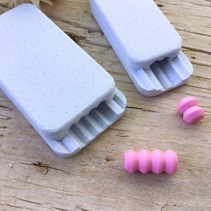 Puede incluir: Dos moldes rectangulares blancos con una superficie texturizada y una serie de crestas en la parte inferior. Dos cuentas de arcilla polimérica rosa están al lado de los moldes.