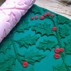 Op de afbeelding: Een roze deegroller met een bladerenpatroon ligt naast een groene ondergrond versierd met hulstbladeren en rode bessen. Het ontwerp is in reliëf, waardoor een textuureffect ontstaat. Ideaal voor bakken en knutselen.