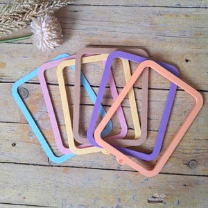 May include: A selection of pastel-coloured rectangular frames in shades of blue, pink, yellow, beige, purple, and peach. The frames are displayed on a wooden surface, with a decorative flower in the upper left corner.
