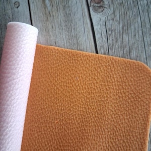 May include: A sheet of textured material with a golden-brown top surface and a rolled edge revealing a pale pink underside. The material features a consistent pebbled grain pattern across the surface, showing a clear contrast between the two colours.