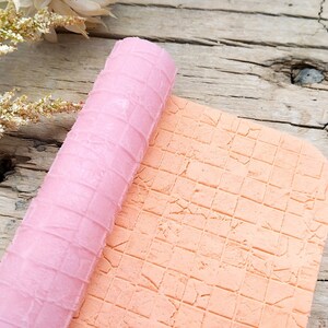 Puede incluir: Un rodillo texturizado rosa y naranja para hornear. El rodillo tiene un patrón cuadrado y está hecho de silicona. La superficie naranja está texturizada con un patrón cuadrado.