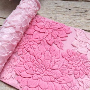 Puede incluir: Un rodillo cilíndrico de color rosa claro con un patrón floral en relieve, que se presiona sobre una lámina plana de material rosa a juego para crear un diseño profundo y grabado de varias flores grandes y superpuestas tipo dalia.