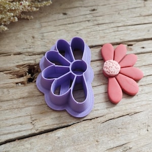 Peut inclure: Un emporte-pièce en forme de marguerite violette à côté d'une marguerite rose finie. Le découpoir est en plastique et présente un design détaillé. La marguerite finie a des pétales roses et un centre texturé.