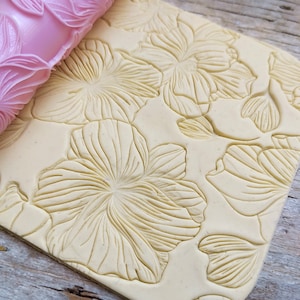 Könnte beinhalten: Ein rosa Nudelholz mit einem Blumenmuster rollt über eine beige Fondant-Platte mit einem geprägten Blumenmuster.