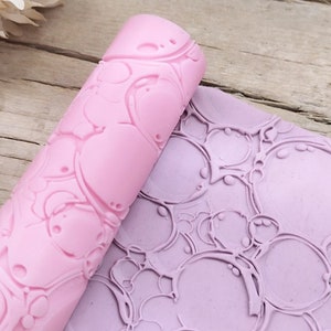 Può includere: Un mattarello in silicone rosa con un motivo a bolle. Il mattarello è appoggiato su un tappetino in silicone viola con lo stesso motivo a bolle.