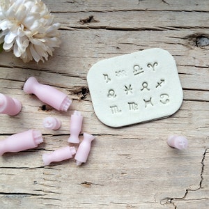 Könnte beinhalten: Ein Set mit 12 rosa Stempeln mit Tierkreiszeichen und einer weißen Plastikfolie, auf der die Tierkreiszeichen abgebildet sind. Die Stempel liegen auf einer Holzoberfläche.