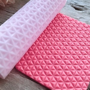 Könnte beinhalten: Eine rosa und weiße Silikon-Nudelholz und -Matte mit einem geometrischen Diamantenmuster. Das Nudelholz liegt auf der Matte.