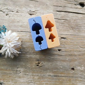 Puede incluir: Molde de silicona de dos piezas para hacer manualidades de resina. El molde está dividido en dos secciones, una azul y una naranja. La sección azul tiene dos cavidades en forma de hongo, mientras que la sección naranja tiene dos cavidades en forma de árbol.