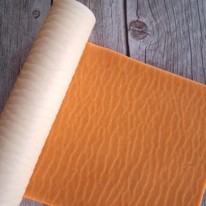 May include: A cylindrical white rolling pin with a textured surface featuring a repeating wave pattern, positioned next to a flat, square orange sheet of material that displays the same embossed wave texture across its entire surface area.