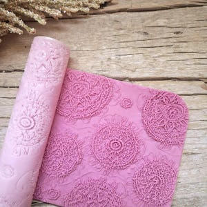 Può includere: Un set rosa di mattarello e tappetino in silicone. Il mattarello è arrotolato, mostrando un motivo floreale dettagliato. Il tappetino presenta un design ripetitivo simile al pizzo. Il set è per la cottura e l'artigianato.