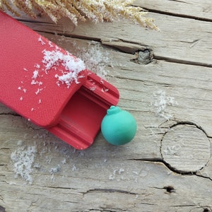 Peut inclure: Un traîneau en plastique rouge avec une balle verte à l'intérieur. Le traîneau est sur une surface en bois avec des flocons blancs ressemblant à de la neige.