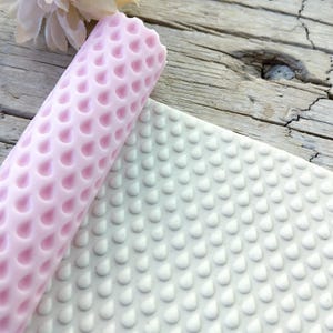 Puede incluir: Un rodillo de silicona rosa con un patrón de lágrima y una estera de silicona blanca con un patrón de lágrima.