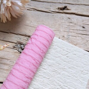 Könnte beinhalten: Eine rosa texturierte Rolle zum Erstellen von Mustern auf Fondant oder Ton. Die Rolle liegt auf einer weißen, strukturierten Oberfläche.