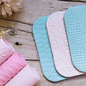 Op de afbeelding: Een verzameling van getextureerde kunstleren vellen in pastelkleuren, waaronder roze, lichtblauw en crème. De vellen hebben reliëfpatronen zoals golven, slangenleer en krokodillenleer. Opgerolde roze vellen zijn zichtbaar op de voorgrond.
