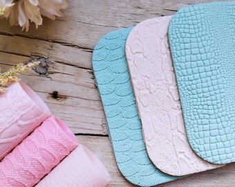 May include: A collection of textured faux leather sheets in pastel colors, including pink, light blue, and cream. The sheets feature embossed patterns like waves, snake scales, and crocodile skin. Rolled pink sheets are visible in the foreground, with a wooden background.