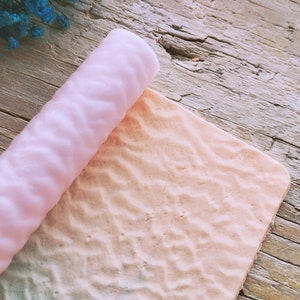 Puede incluir: Un rodillo de silicona rosa con una superficie texturizada. El rodillo se está extendiendo sobre una superficie texturizada rosa claro.
