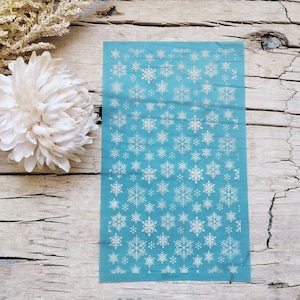 May include: A sheet of white snowflake nail decals on a light blue background. The snowflakes are various sizes and shapes.