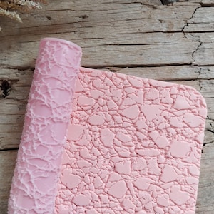 Könnte beinhalten: Eine hellrosa zylindrische Teigrolle mit einer strukturierten Oberfläche aus unregelmäßigen, rissigen Formen. Neben der Rolle liegt ein flaches, quadratisches Stück hellrosa Material mit demselben geprägten Muster aus fragmentierten Formen.