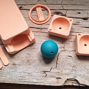 Può includere: Oggetti stampati in 3D color pesca, tra cui una custodia rettangolare, un anello e due contenitori quadrati con interni arrotondati. È presente anche una sfera blu acqua con un foro. Gli oggetti sono disposti su una superficie di legno invecchiato.