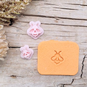 May include: Two pink plastic bee-shaped cookie cutters and an orange silicone baking mat with a bee design embossed on it.