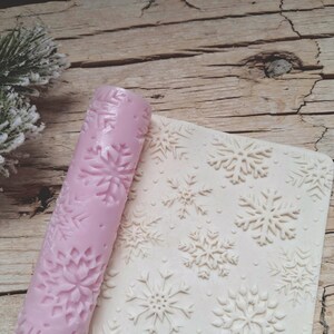 May include: A pink rolling pin and a textured mat, both featuring snowflake patterns. The rolling pin is cylindrical, and the mat is rectangular with raised snowflake designs. The items are likely for baking or crafting.