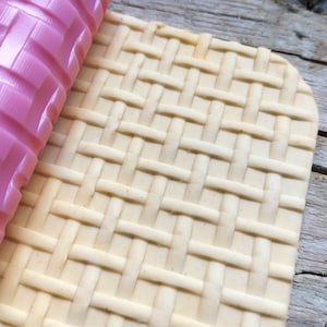 Puede incluir: Un rodillo de fondant blanco con un patrón tejido. El rodillo se utiliza para crear un diseño texturizado en una pieza de fondant.