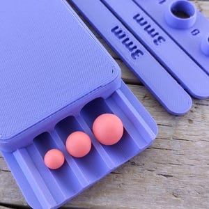 Puede incluir: Un juego de herramientas de artesanía de color lavanda con una bandeja que contiene tres esferas rosas de diferentes tamaños. El juego incluye herramientas de medición con marcas de 3 mm. Las herramientas están sobre una superficie de madera envejecida.