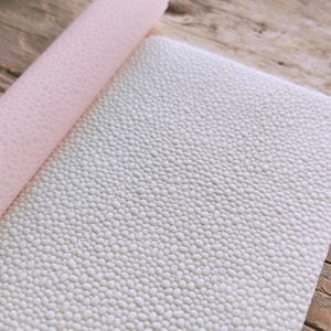 Peut inclure: Tapis de cuisson en silicone blanc avec un motif texturé de galets. Le tapis est enroulé et partiellement déroulé, révélant la surface texturée.