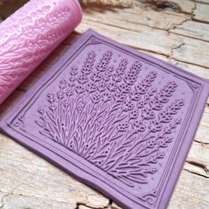 Könnte beinhalten: Ein hellrosa zylindrisches Nudelholz und ein quadratischer violetter Stempel, beide mit einem erhabenen, detaillierten Prägemuster aus Lavendelzweigen. Der Stempel hat einen dünnen Rahmen, und das Nudelholz zeigt ein florales Muster.
