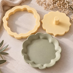 May include: A set of three scalloped-edge items including a pale yellow circular frame, a matching pale yellow solid circular press with a central handle, and a sage green circular tray with a matching scalloped rim and recessed center.