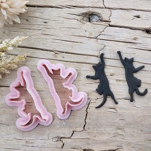 May include: Two pink plastic cookie cutters shaped like cats in a jumping pose. The cutters are on a wooden surface.