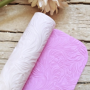 Könnte beinhalten: Eine weiße und eine rosa Silikon-Nudelholz und -matte mit einem wirbelnden Blatt-Muster. Das Nudelholz ist aufgerollt und die Matte liegt flach.