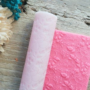 Puede incluir: Un rodillo de silicona rosa y una lámina de silicona rosa con un diseño floral. Ambos se utilizan para decorar pasteles y postres.