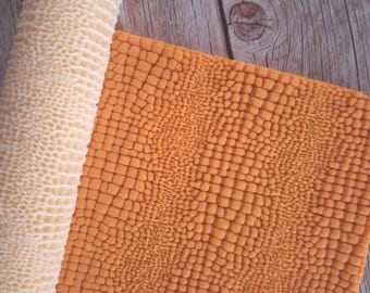 May include: A cylindrical white rolling pin with a deep honeycomb texture pattern positioned next to a flat, rectangular orange sheet of material featuring a raised, grid-like square texture that mimics a reptile skin pattern.