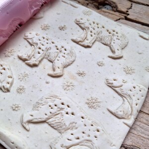 May include: A light-colored, textured rolling pin with a bear and snowflake pattern. The pattern is embossed on a light-colored surface, with a pink roller visible in the upper left corner. The design features multiple bear figures and snowflake shapes.