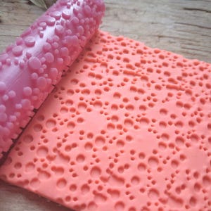 May include: A pink textured rolling pin next to a sheet of pink material with a matching pattern. The rolling pin has a series of raised, rounded shapes. The sheet has a pattern of holes and raised areas, creating a textured surface.