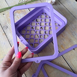 May include: A collection of lavender-coloured, square craft stencils with rounded corners. The stencils feature a grid pattern in the centre and are labelled with numbers. The stencils are stacked, with one stencil slightly removed, showing the layers.