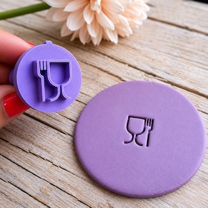 Puede incluir: Un sello circular morado con un diseño de tenedor y copa de vino. El sello lo sostiene una mano con esmalte de uñas rojo. Un círculo morado a juego con el mismo diseño está al lado del sello. Una flor melocotón está en el fondo.