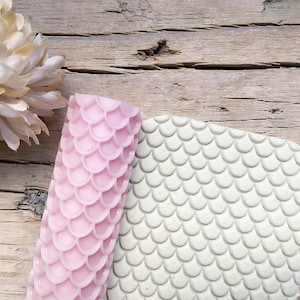 Può includere: Un mattarello cilindrico rosa con un motivo a squame di pesce in rilievo e un pezzo piatto di argilla verde chiaro con un'identica texture impressa, che mostra il risultato dello strumento utilizzato per creare il motivo ripetuto.