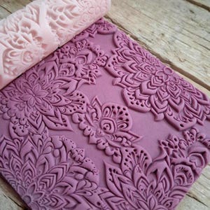 Könnte beinhalten: Eine rosa Teigrolle mit floralem Muster liegt auf einem lila Teigblatt, das ebenfalls mit dem gleichen Design bedruckt ist. Die Teigrolle und der Teig erzeugen einen strukturierten, dekorativen Effekt, ideal zum Backen und Basteln.